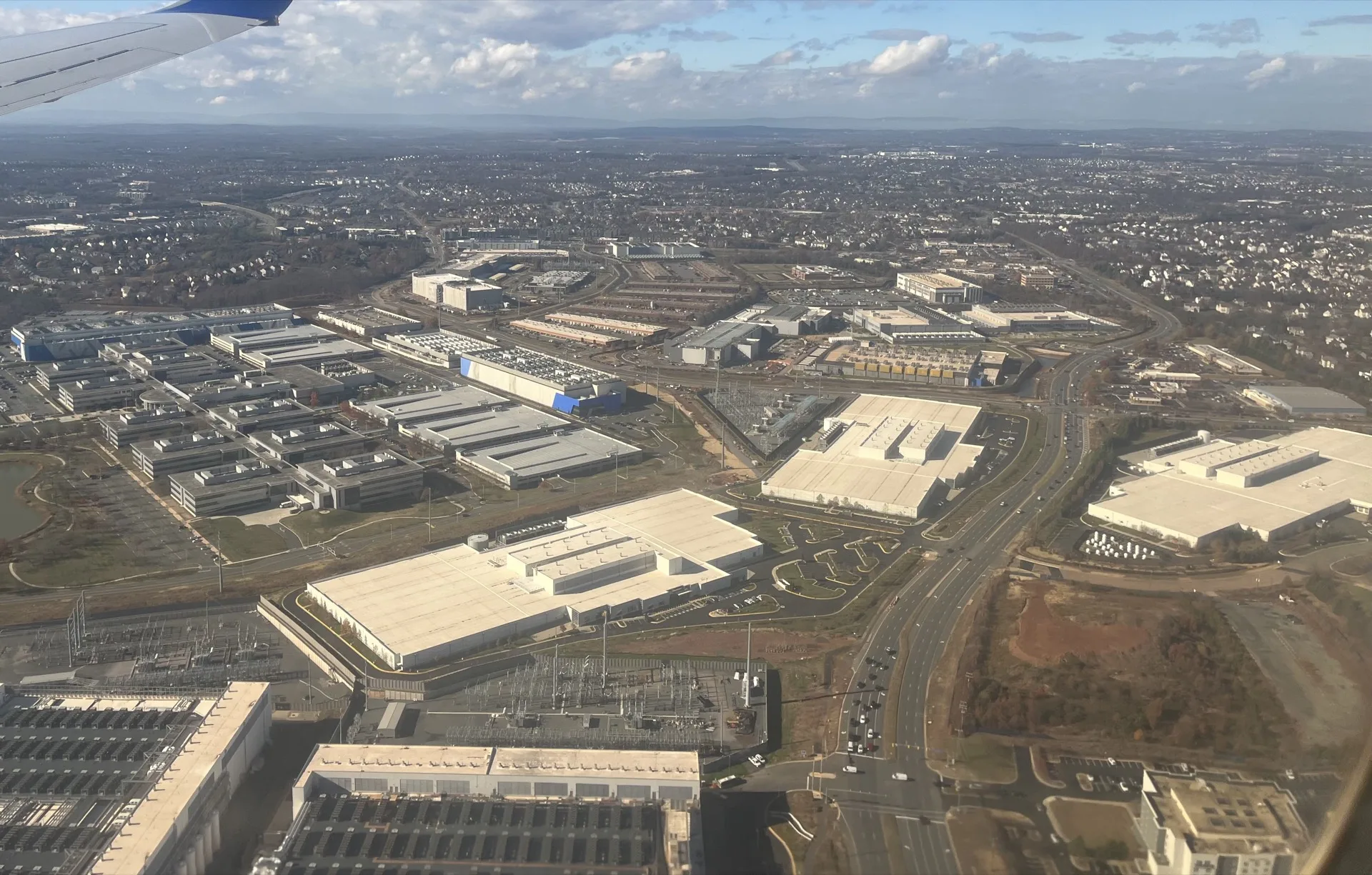 Aerial view from an airplane window of a dense cluster of data centers and warehouses in Ashburn, Virginia, with residential neighborhoods stretching to the horizon behind them.