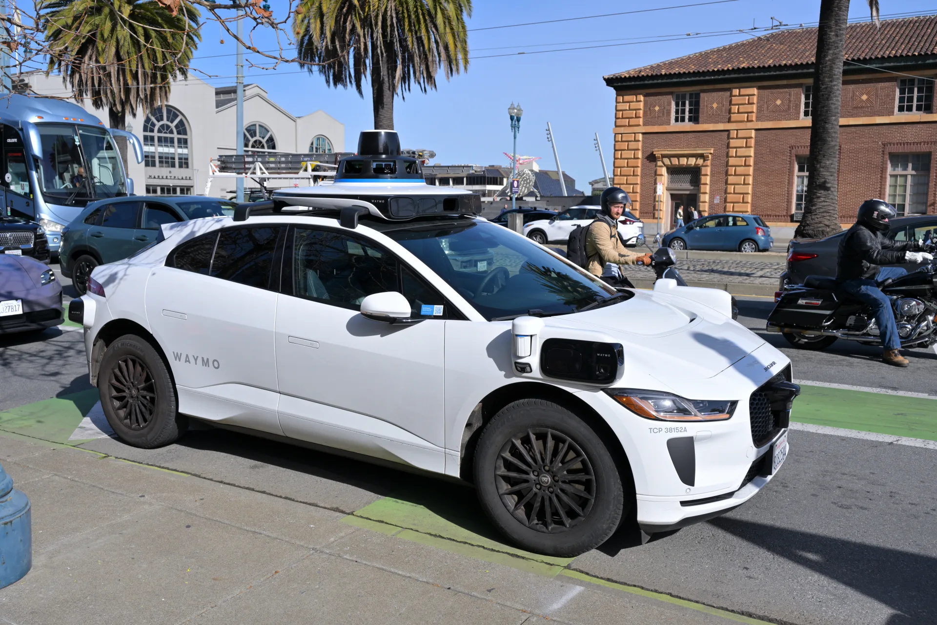 A white Waymo self-driving Jaguar taxi stopped in a green bike lane on San Francisco's Embarcadero, a motorcyclist passing close on the right.